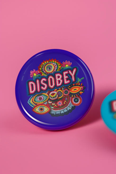 Stay Weird" black pinback button with pearl-effect lettering surrounded by folk art flowers in red and turquoise on a dark background.